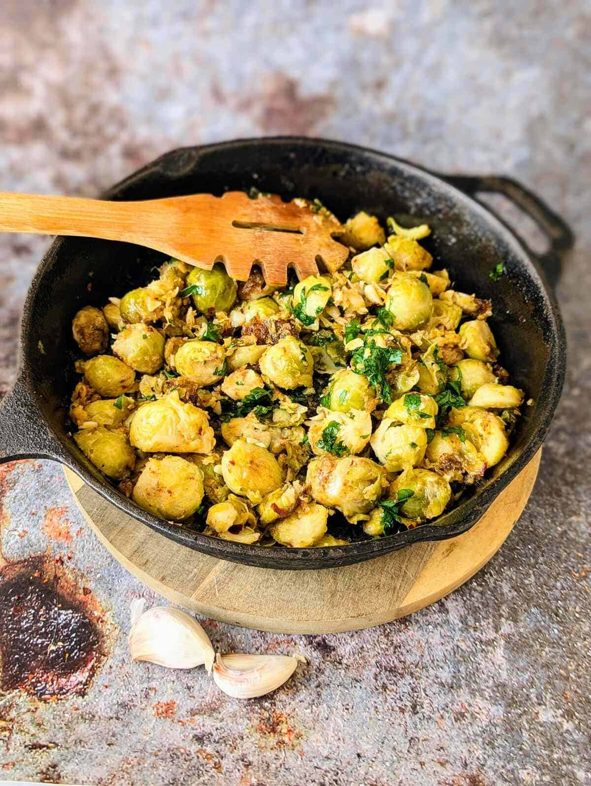 A skillet filled with Brussels sprouts and herbs. A wooden spoon rests on the pan. Two garlic cloves lie infront.