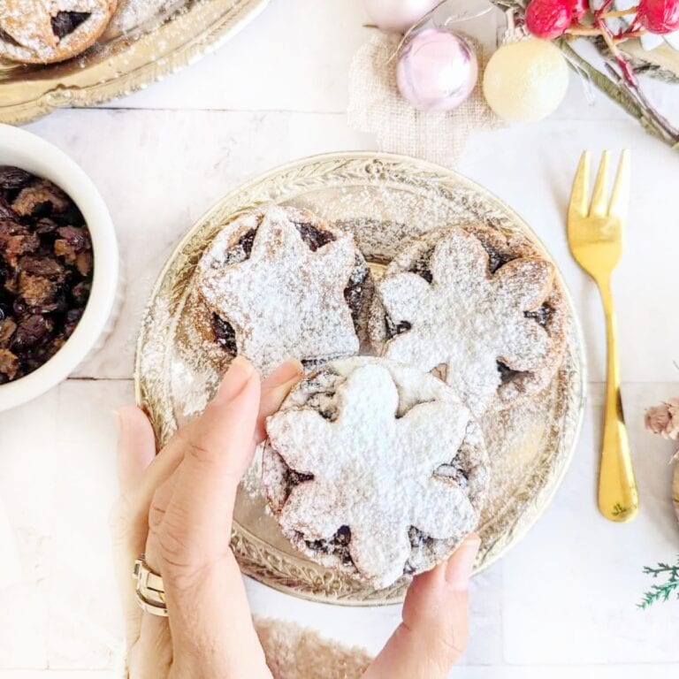 A hand holding a vegan mince pie. Christmas decorations and a gold fork in the background.