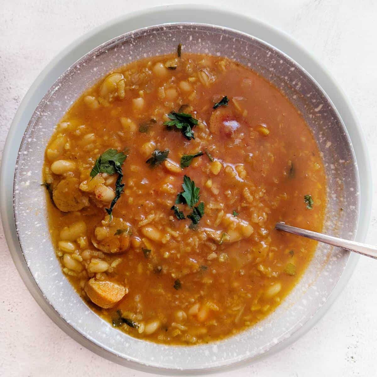 A dish of tofy sausage bean and rice soup. A spoon is in the soup and a plate underneath.