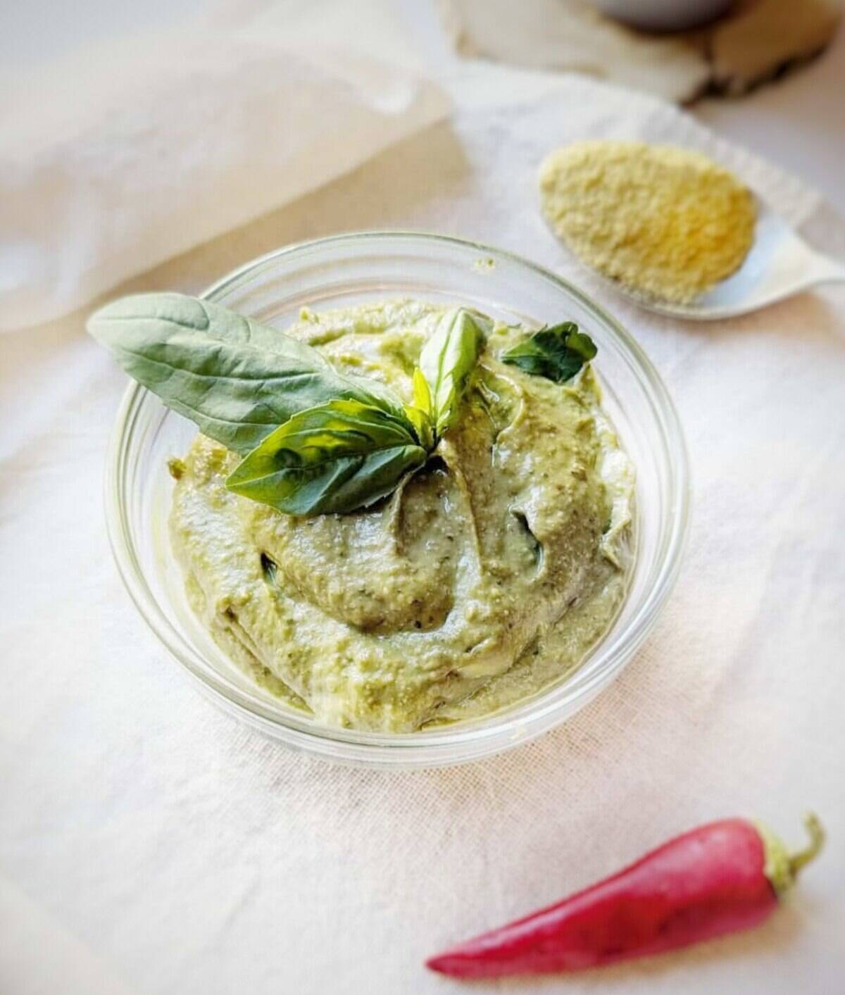 Fresh avocado chilli pesto garnished with basil leaves, served in a glass bowl. A chilli pepper is lying infront of the bowl.