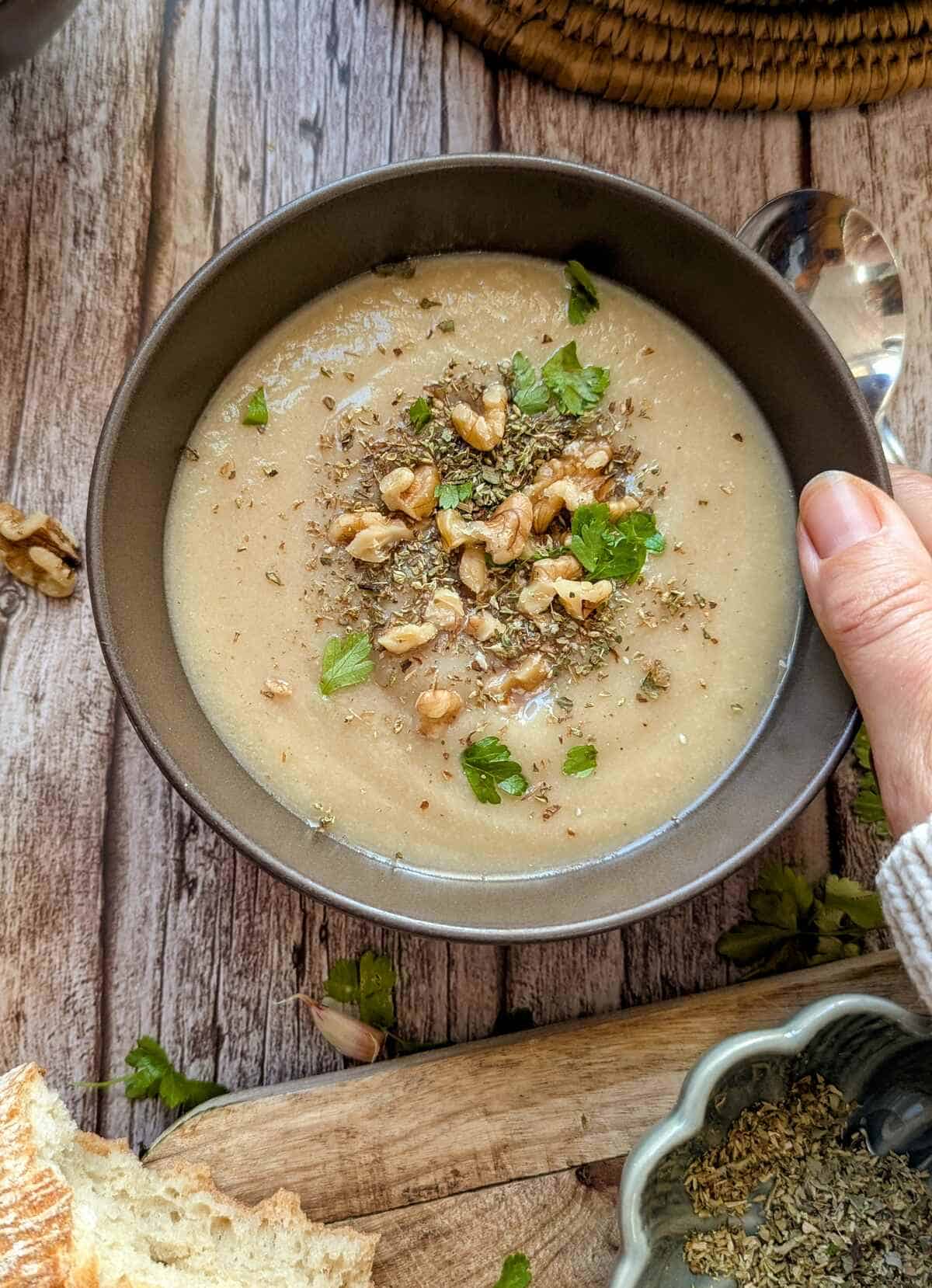 A hand holding the bowl of potato cauliflower soup. Bread and a bowl of oregano are infront.