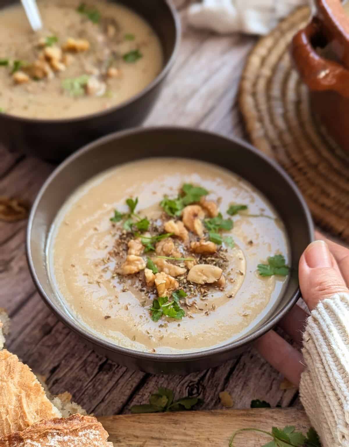 A hand holding a cozy bowl of creamy potato cauliflower soup. A faded bowl is in the background.