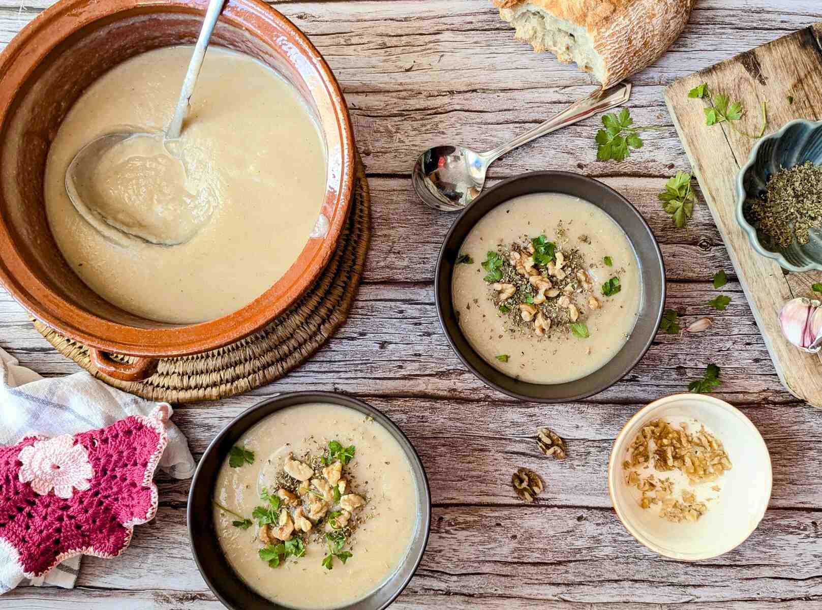 Three bowls of creamy potato cauliflower soup in a bowl with bread, a sppon, walnuts and oregano near.