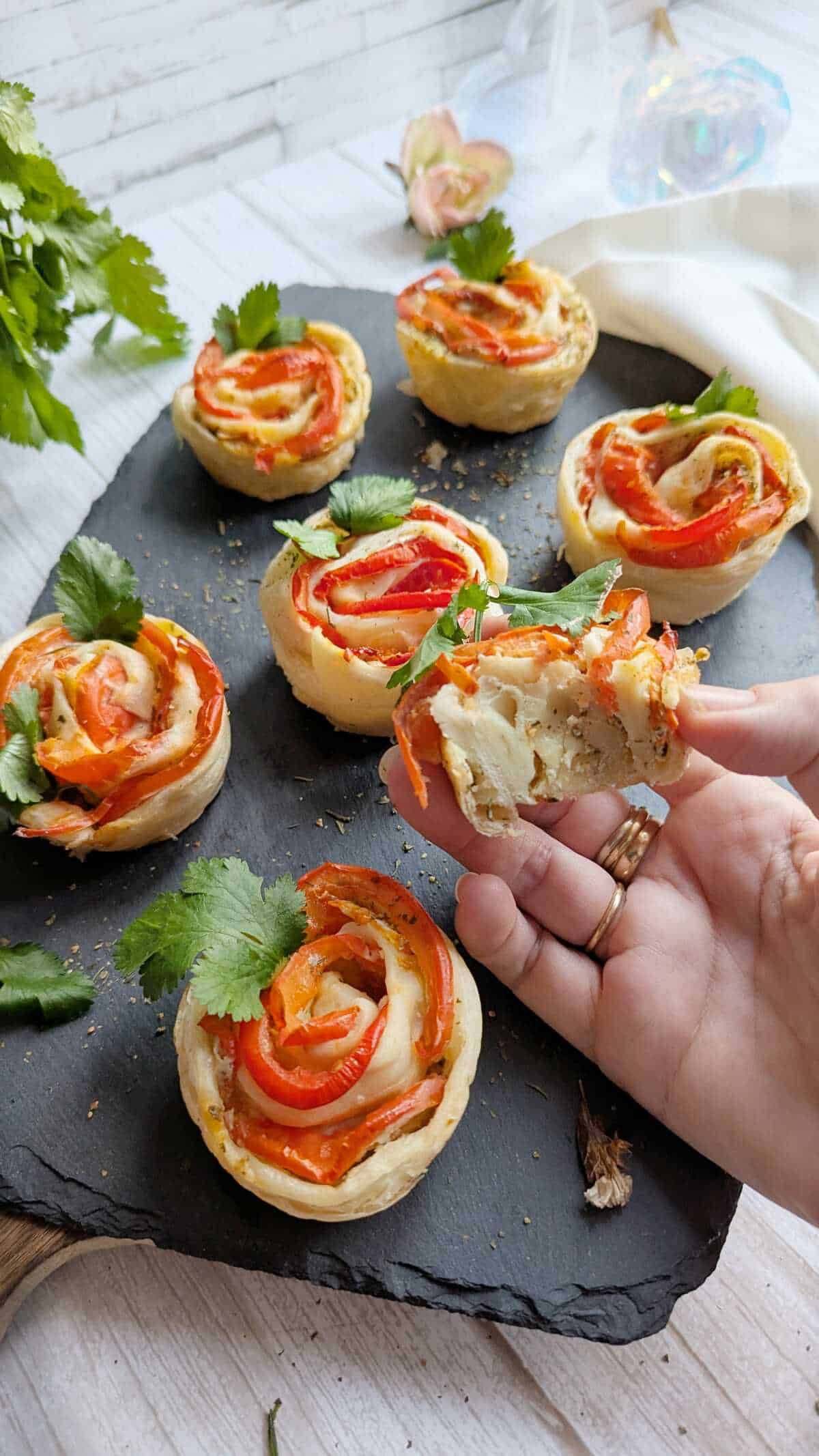 Puff pastry tomato roses on a black tray. A hand is holding one that is half eaten.