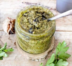 A jar of black garlic vegan pesto with a spoon dipped in and black garlic cloves are by the side.