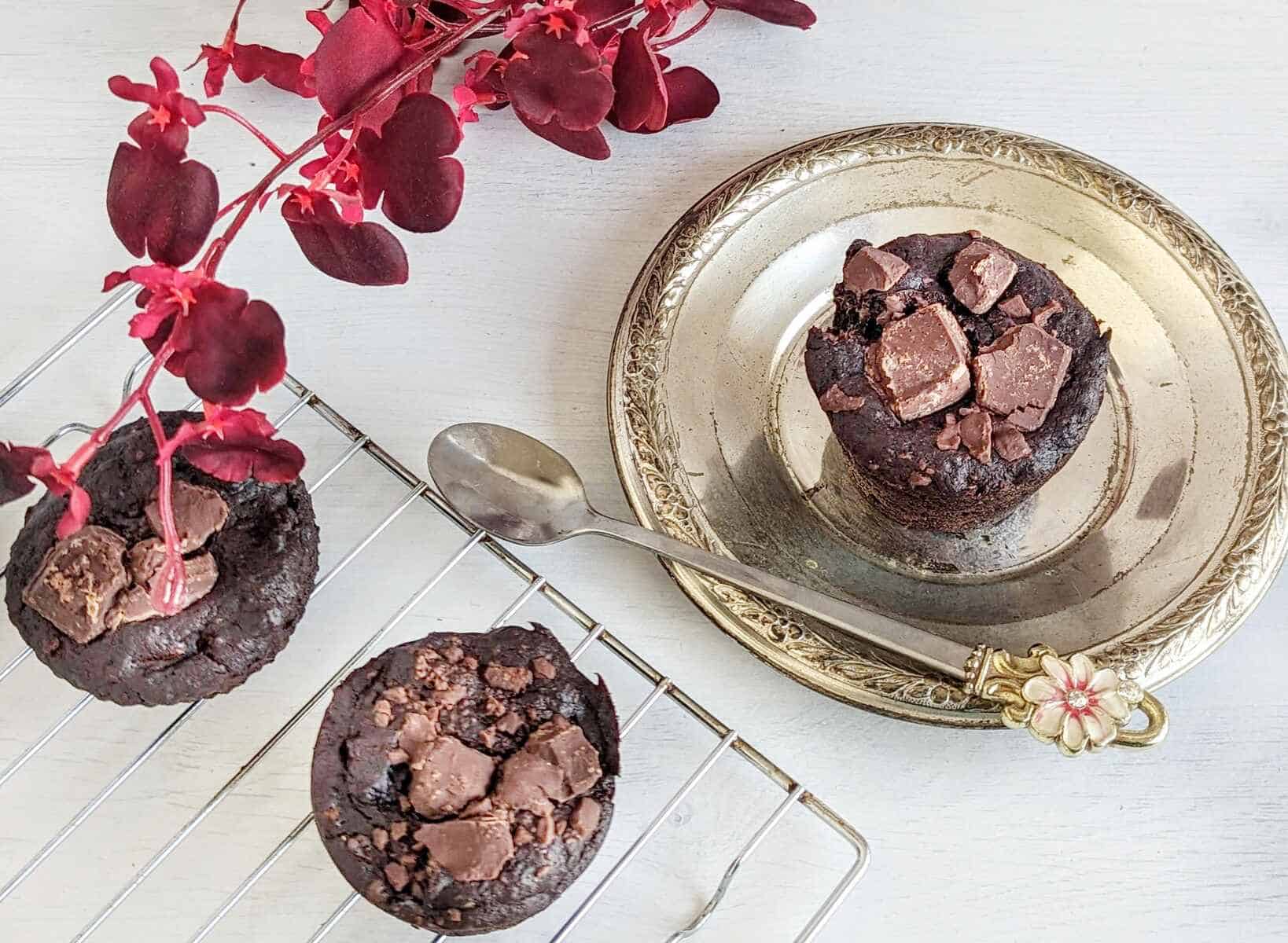 Vegan chocolate banana muffins with chunky topping on a cooling rack and plate. Red flowers are faded in the background.