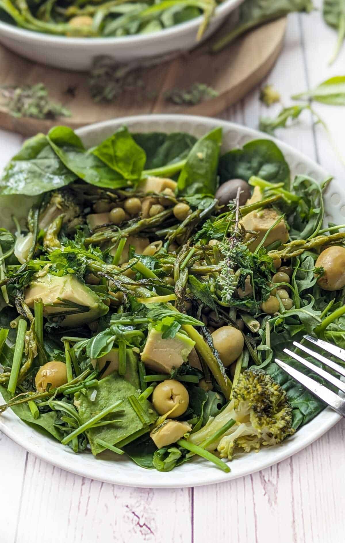 A plate of assorted fresh vegetables and herbs including spinach, avocado, peas, and broccoli. A fork is by the side.