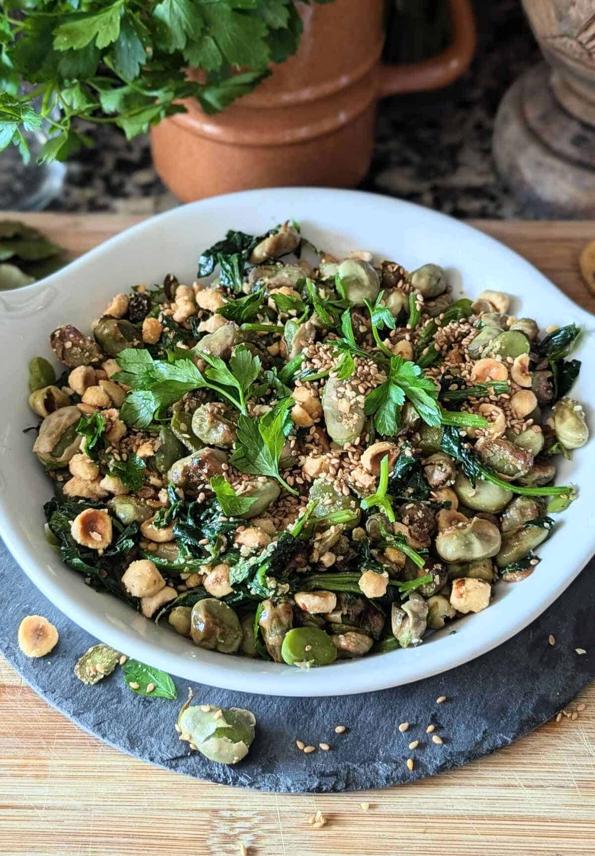 A dish of sautéed spinach and fava bean, with a rustic jug and parsley at the back.