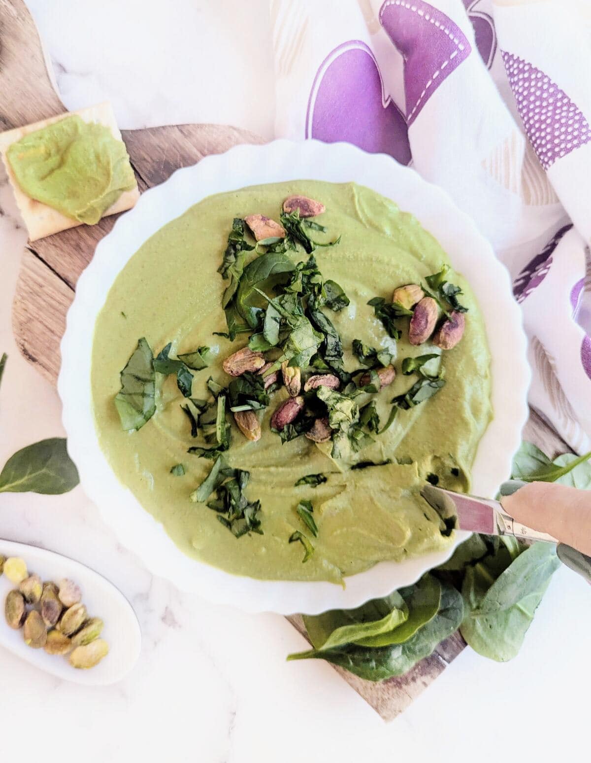 A creamy bowl of spinach and pistachio hummus. Chopped spinach decorated on top and pistachios by the side.