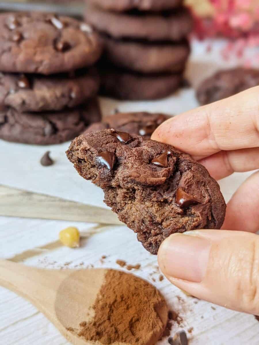 Flourless chocolate cookies with chunks of chocolate. A hand holding a half-eaten cookie. A spoon of cinnamon is sitting in front of the image.